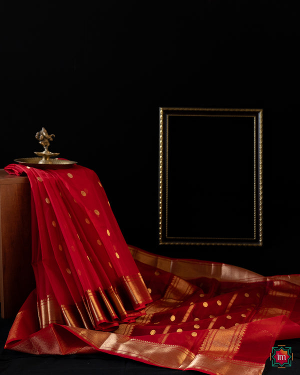 Beautiful red silk saree is pleated and displayed upon a bench with a ganapati statue upon it and with a black background.