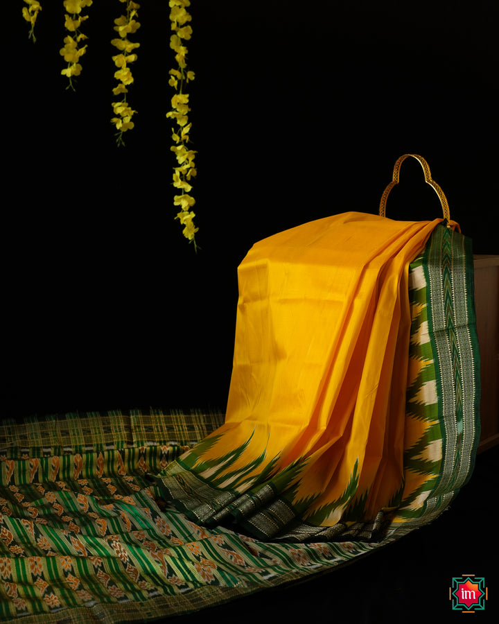 Elegant Yellow Green silk mark saree is displayed on the floor with black background and yellow flowers hanging above.