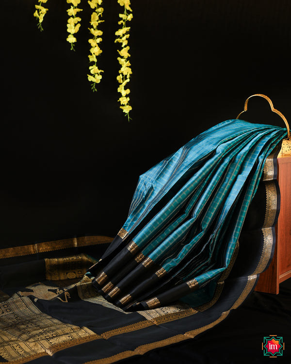 Elegant pacific blue kanjivaram silk saree is displayed on the floor with black background and yellow flowers hanging above.