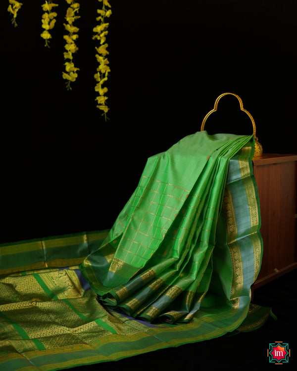 Beautiful Rexona Green Kanjivaram Saree is displayed on the floor with black background and yellow flowers hanging above.