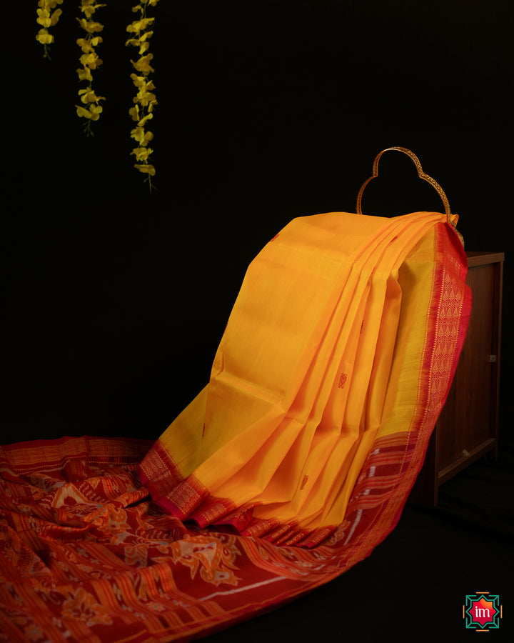 Elegant Yellow Red Khandua Silk Saree Kalyani is displayed on the floor with black background and yellow flowers hanging above.